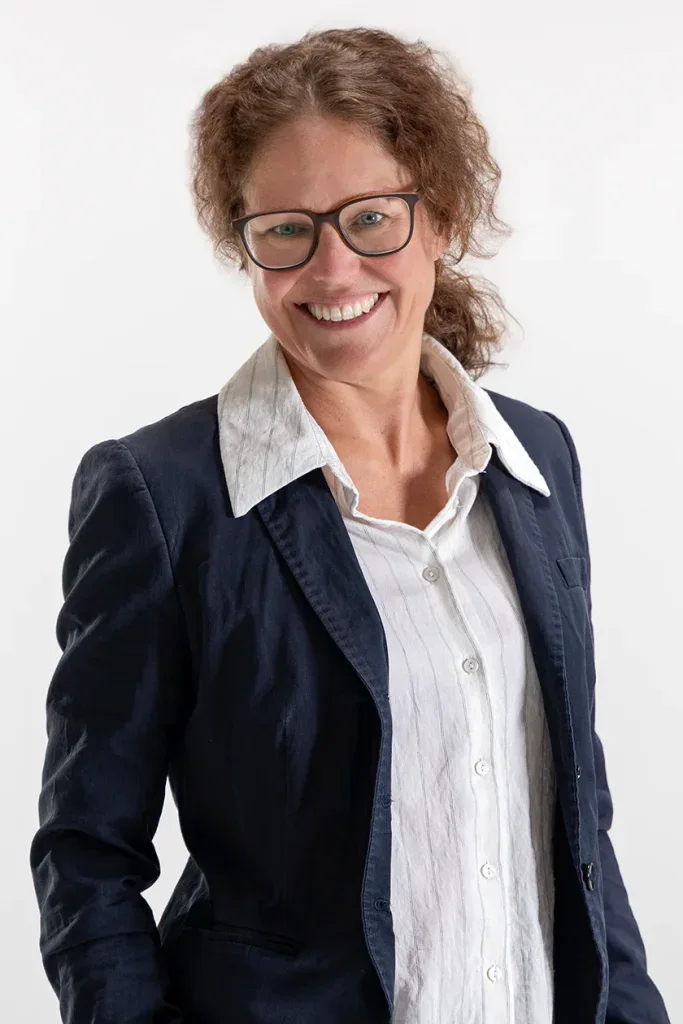 Natürliches Bewerbungsfoto einer lächelnden Frau mit Brille, weisser Bluse und dunklem Blazer vor hellem Hintergrund, aufgenommen im Fotostudio Fokus.