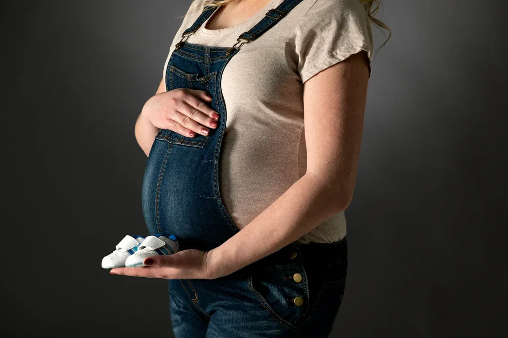 Detailaufnahme beim Babybauch Shooting mit kleinen Babyschuhen, Jeanslatzhose und ruhiger Lichtstimmung