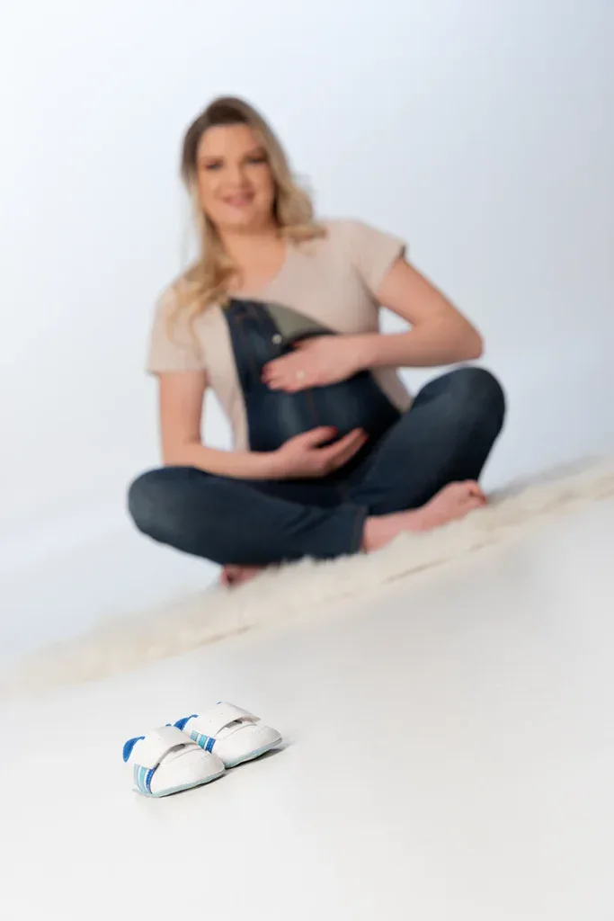 Detailaufnahme beim Babybauch Shooting mit kleinen Babyschuhen, Jeanslatzhose und ruhiger Lichtstimmung