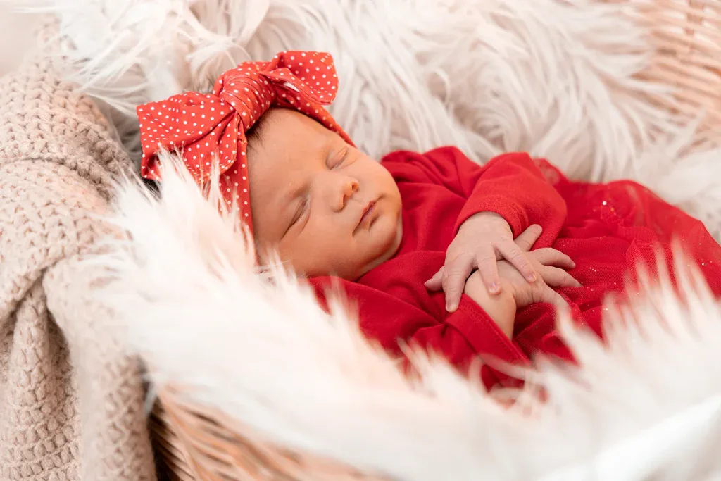 Liebevolles Newborn Foto mit schlafendem Baby in rotem Outfit, grosser Schleife und weicher Decke