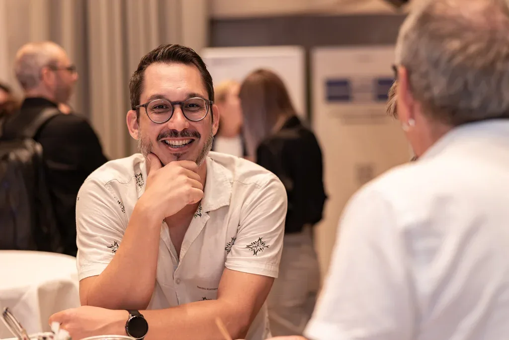 Lachender Teilnehmer sitzt im Gespräch mit einem weiteren Gast an einem Business-Event.