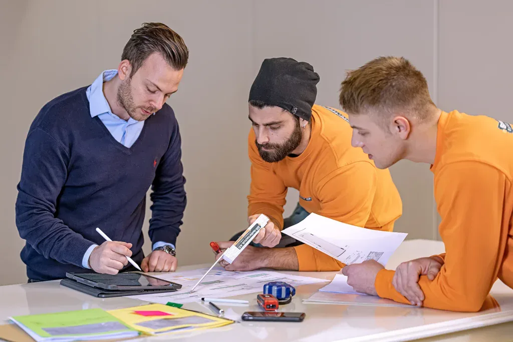 Drei Mitarbeitende besprechen technische Pläne, Werkzeuge und Masse an einem Arbeitstisch.