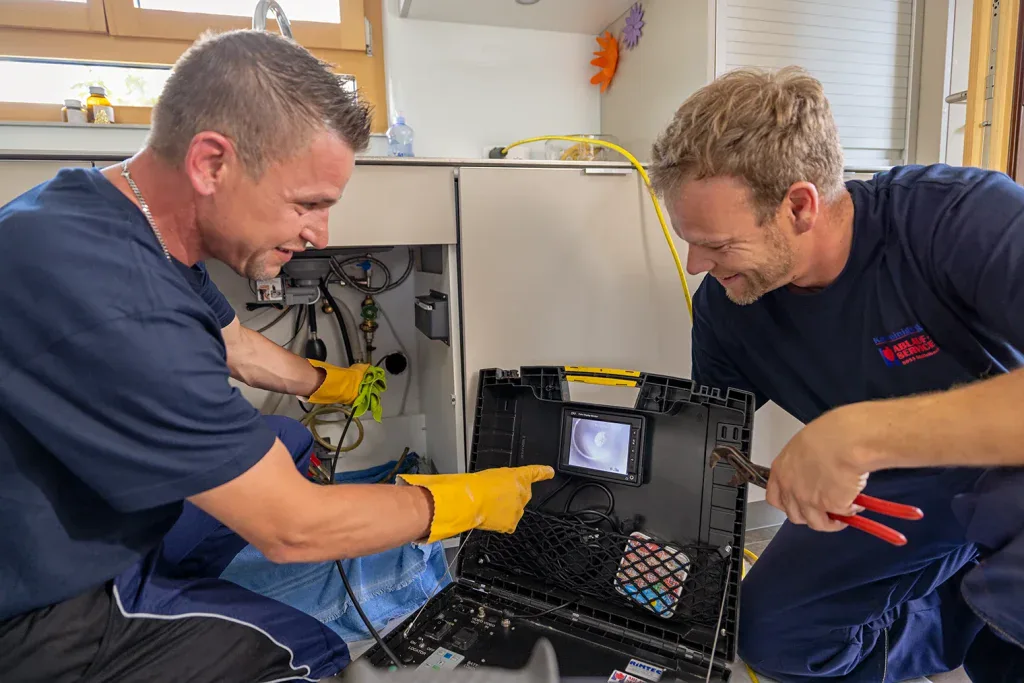 Zwei Servicetechniker prüfen mit Werkzeug und Kamerabild eine technische Anlage während eines Serviceeinsatzes.