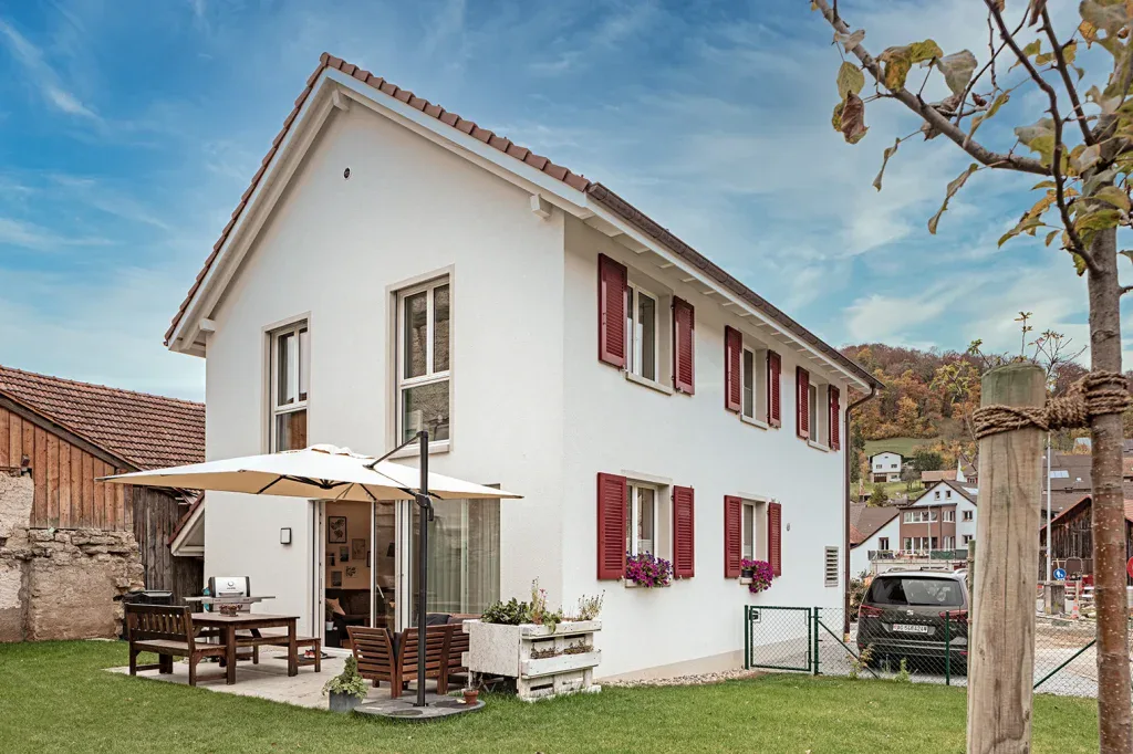 Aussenansicht eines weissen Einfamilienhauses mit rotem Fensterladen und gedecktem Gartensitzplatz.