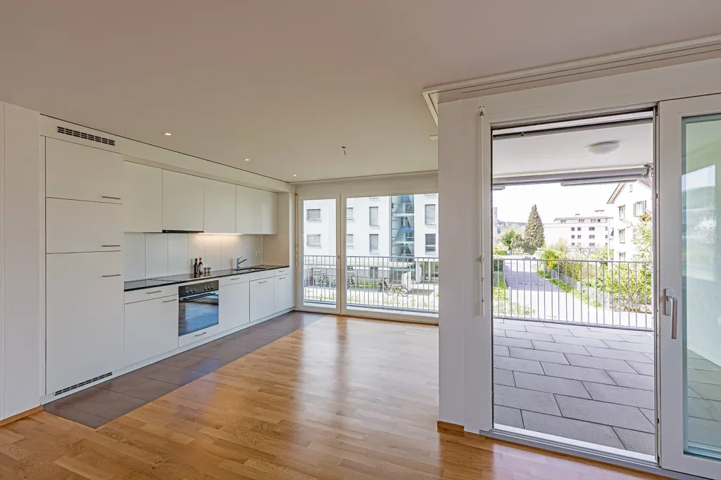 Heller Wohnraum einer modernen Wohnung mit offener Küche, Parkettboden und Zugang zum Balkon.