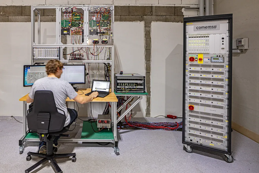 Fachperson arbeitet an mehreren Monitoren neben Schaltschränken und elektronischer Prüftechnik.