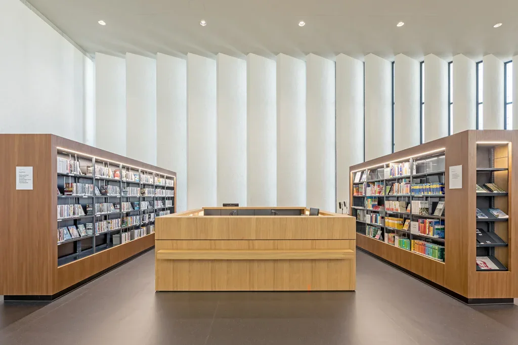Heller Bibliotheksraum mit Bücherregalen, zentralem Holzmobiliar und vertikalen Fensterlamellen in Menzingen.