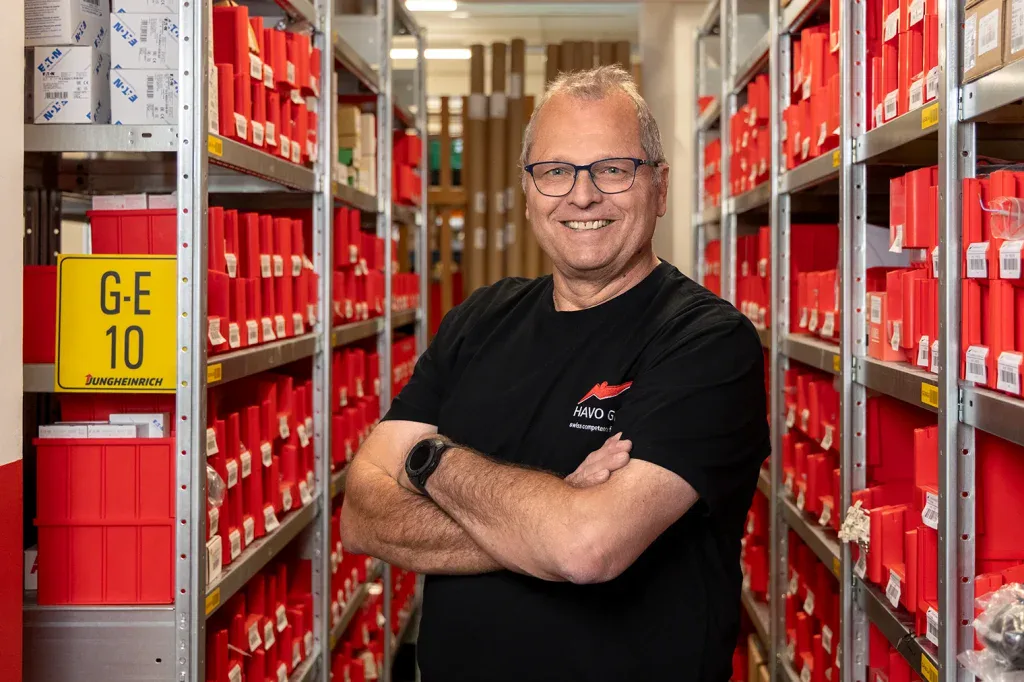 Portrait eines Mitarbeiters vor roten Lagersystemen für Unternehmenswebsite, Mitarbeiterseite und Arbeitgebermarke.