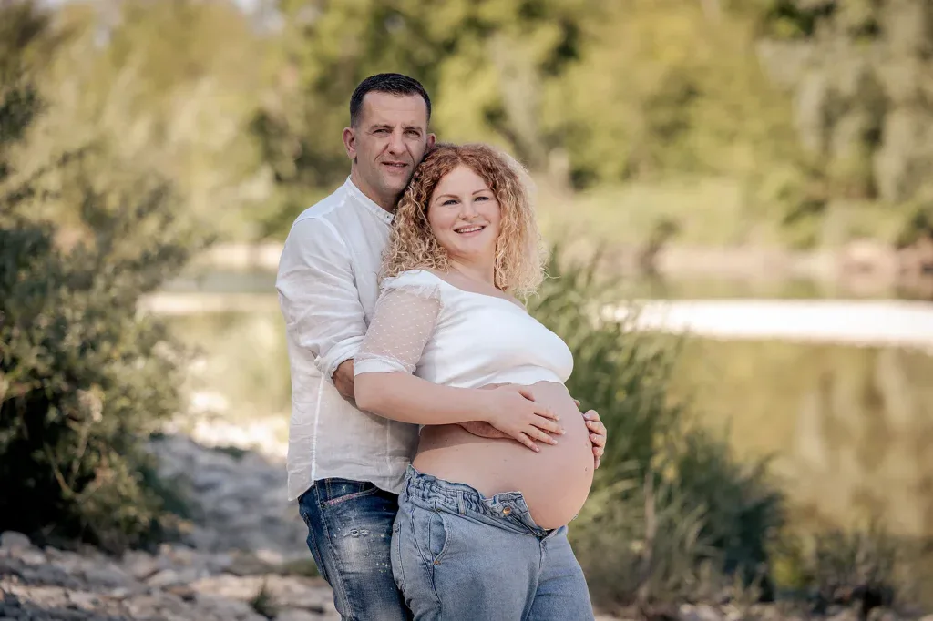Paar bei einem natürlichen Babybauch-Fotoshooting draussen in der Nähe eines Sees.